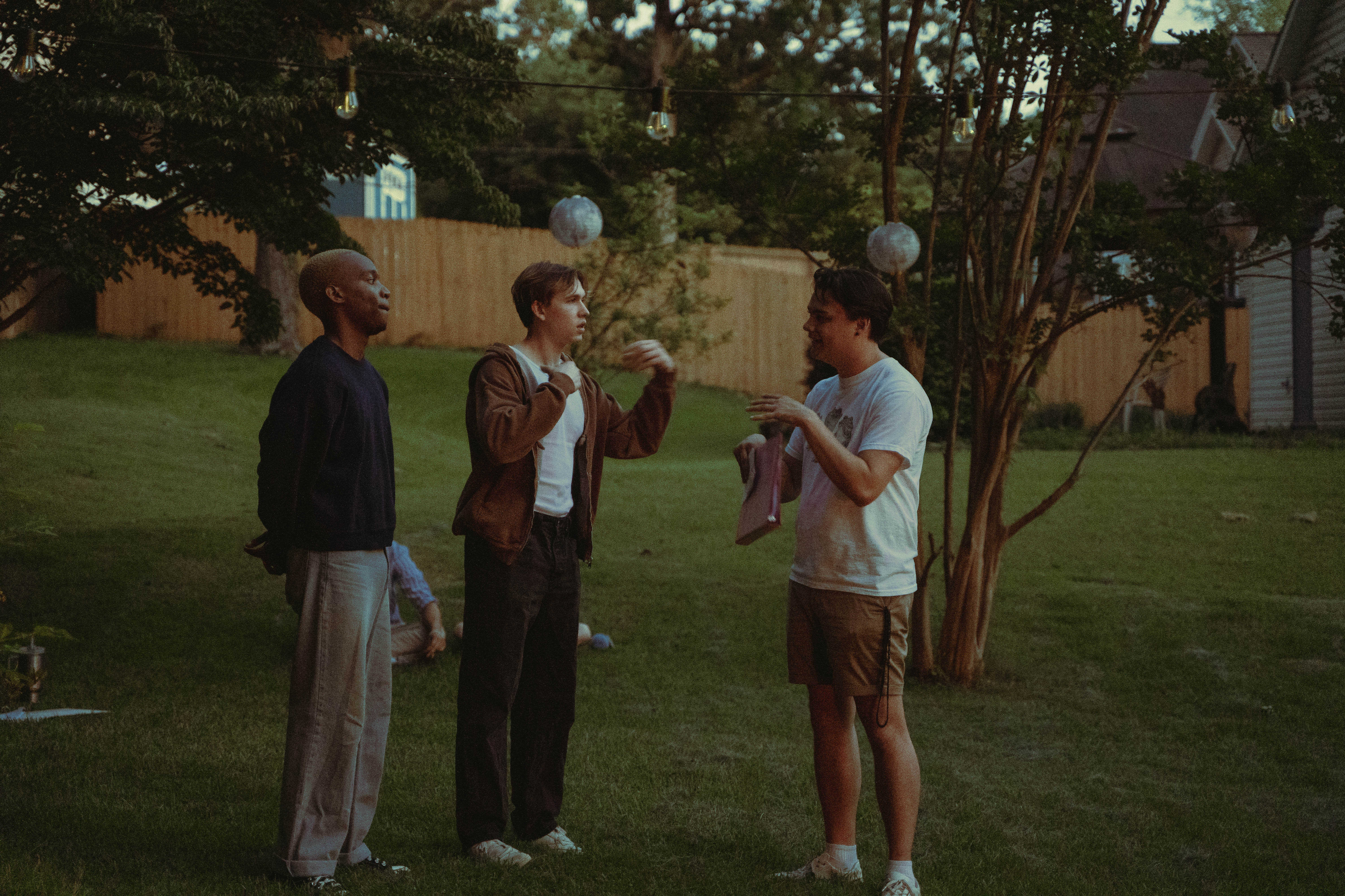 Three young men stand in a grassy backyard at dusk, engaged in conversation under string lights.