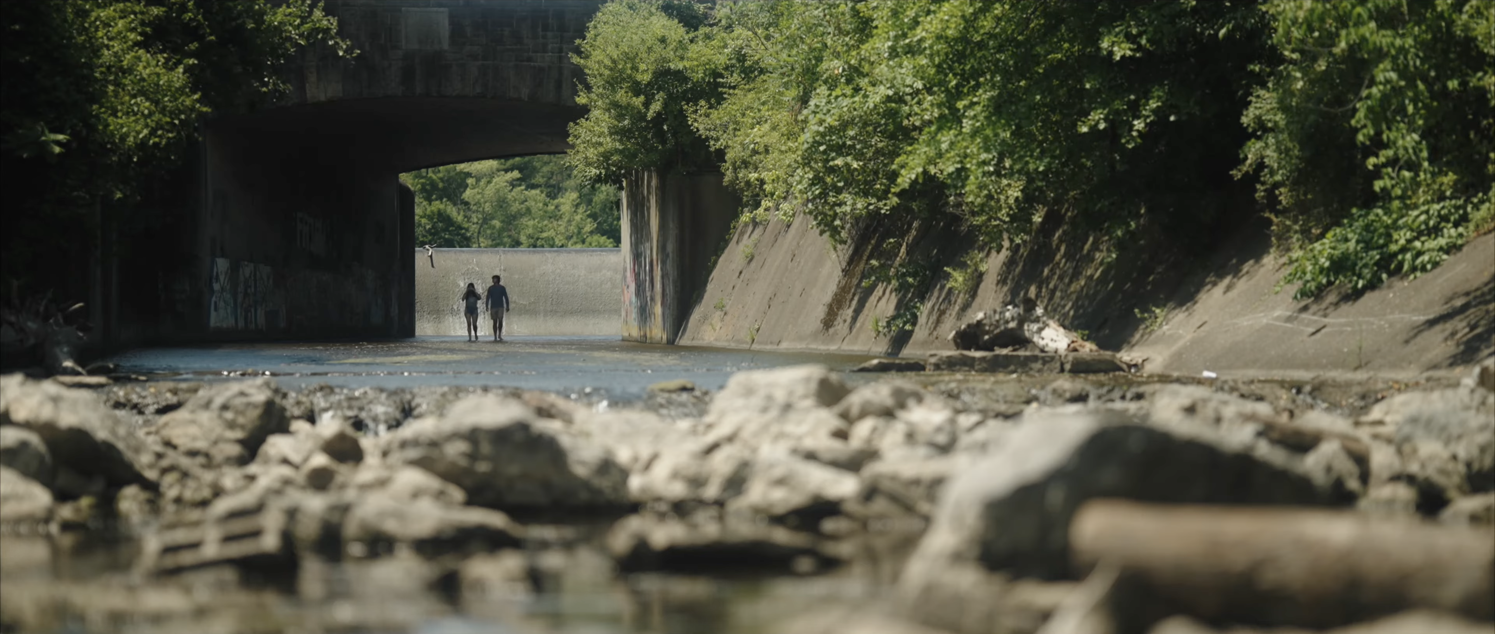 A couple walks through a shallow, rocky riverbed under a concrete bridge towards a small dam.