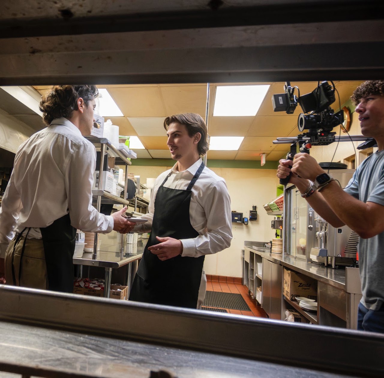 A camera operator films two restaurant staff members in white shirts and black aprons interacting in a kitchen.