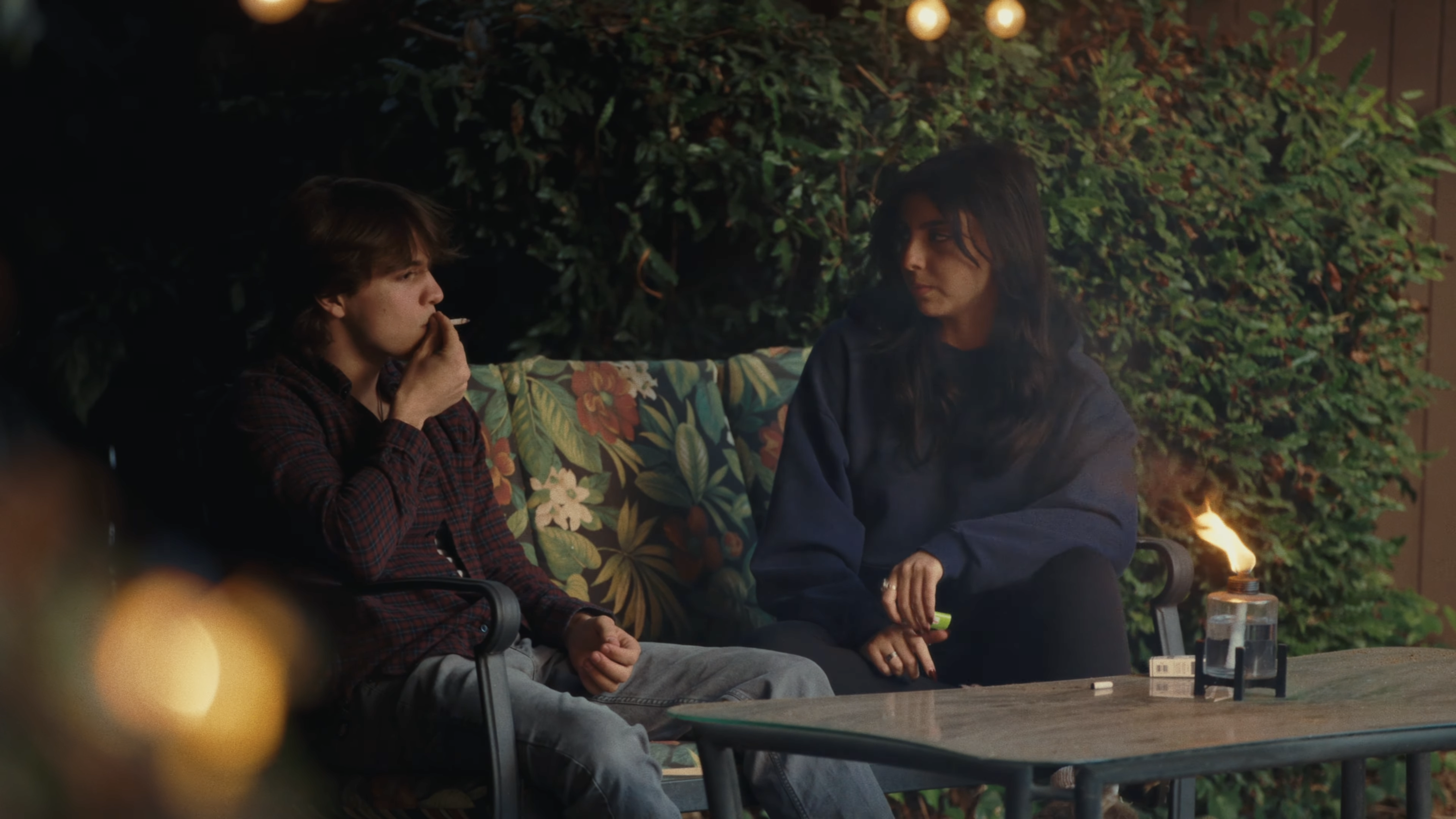 A young man smoking and a young woman sitting together on a floral-patterned outdoor couch at night, with a small lit oil lamp on a glass table in front of them.