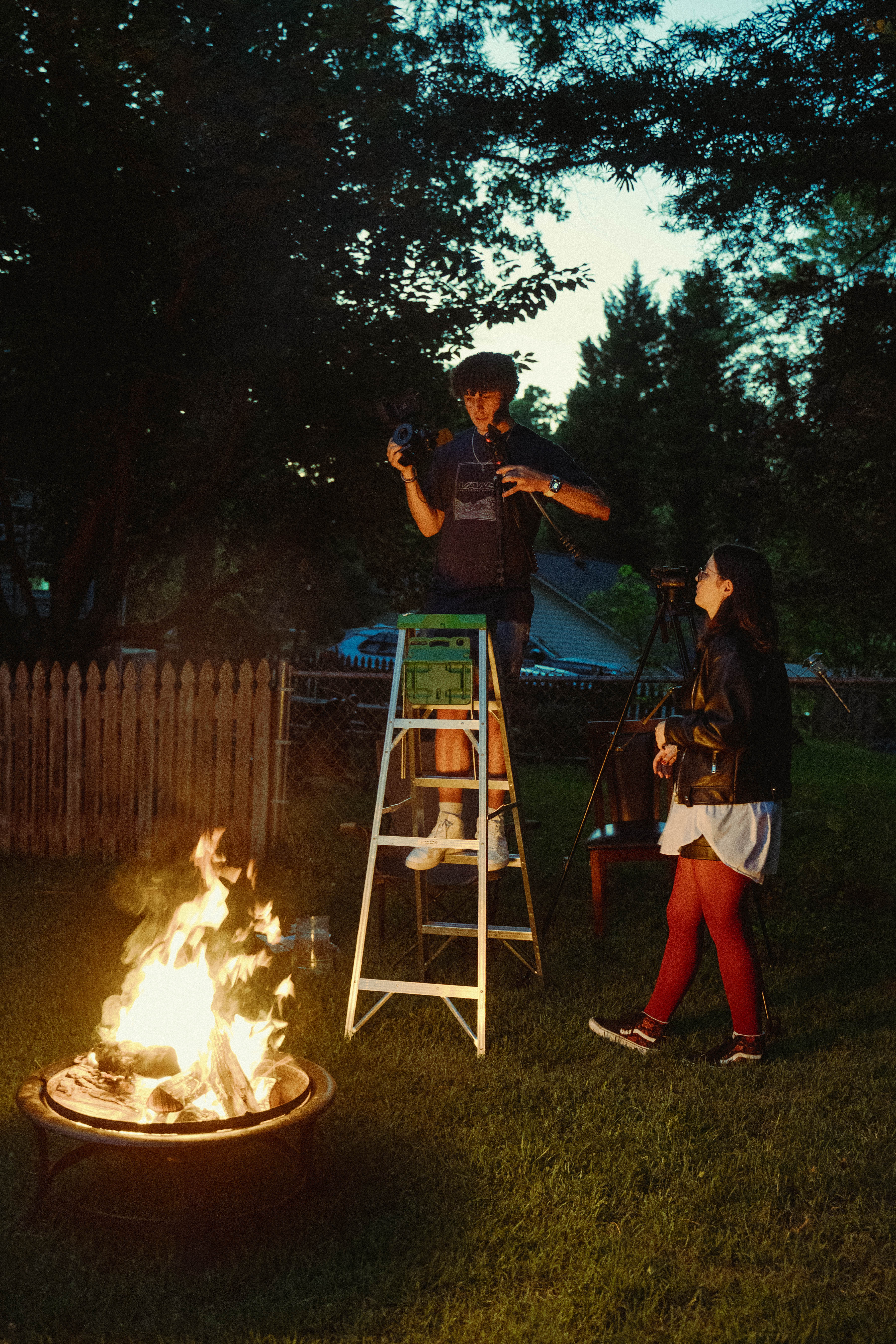 A cinematographer stands on a ladder filming a scene in a backyard at dusk, while a woman stands nearby next to a tripod.