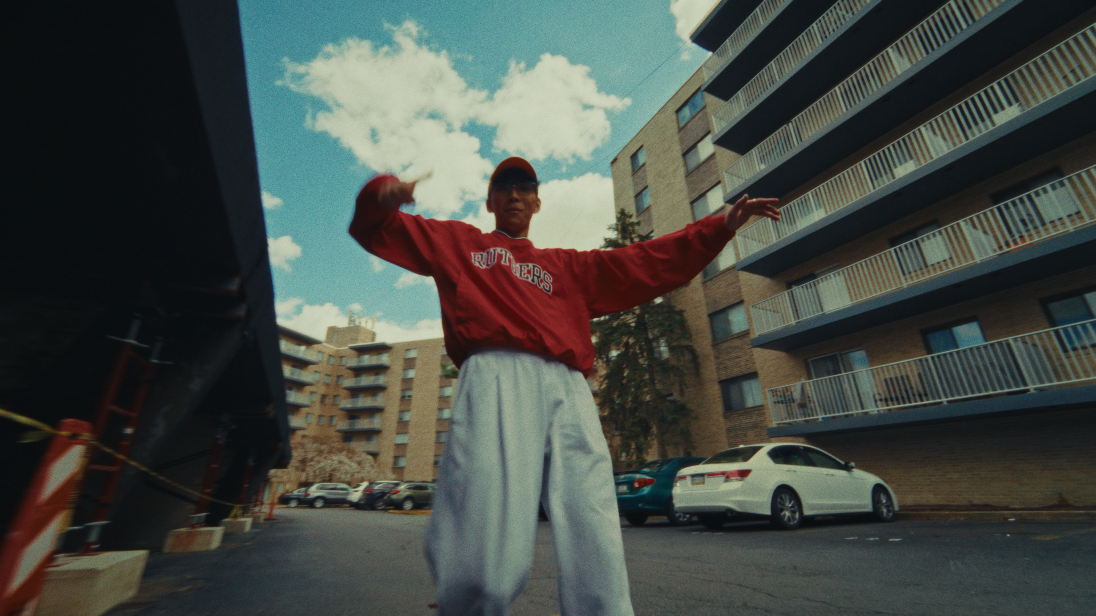 Low-angle view of a young man in a red sweatshirt moving between apartment buildings under a blue sky.