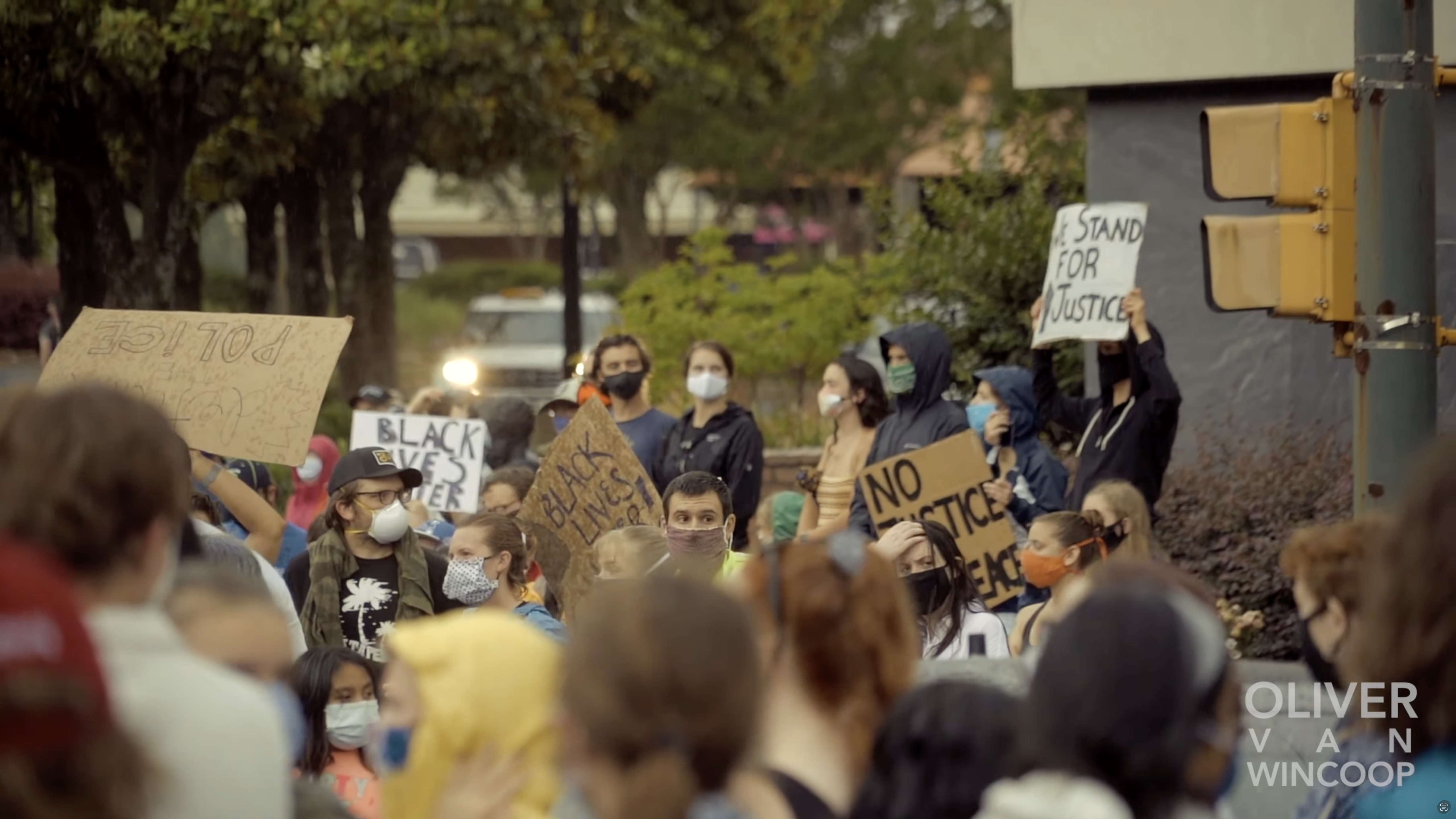 A crowd of people wearing face masks holds protest signs in an outdoor setting.