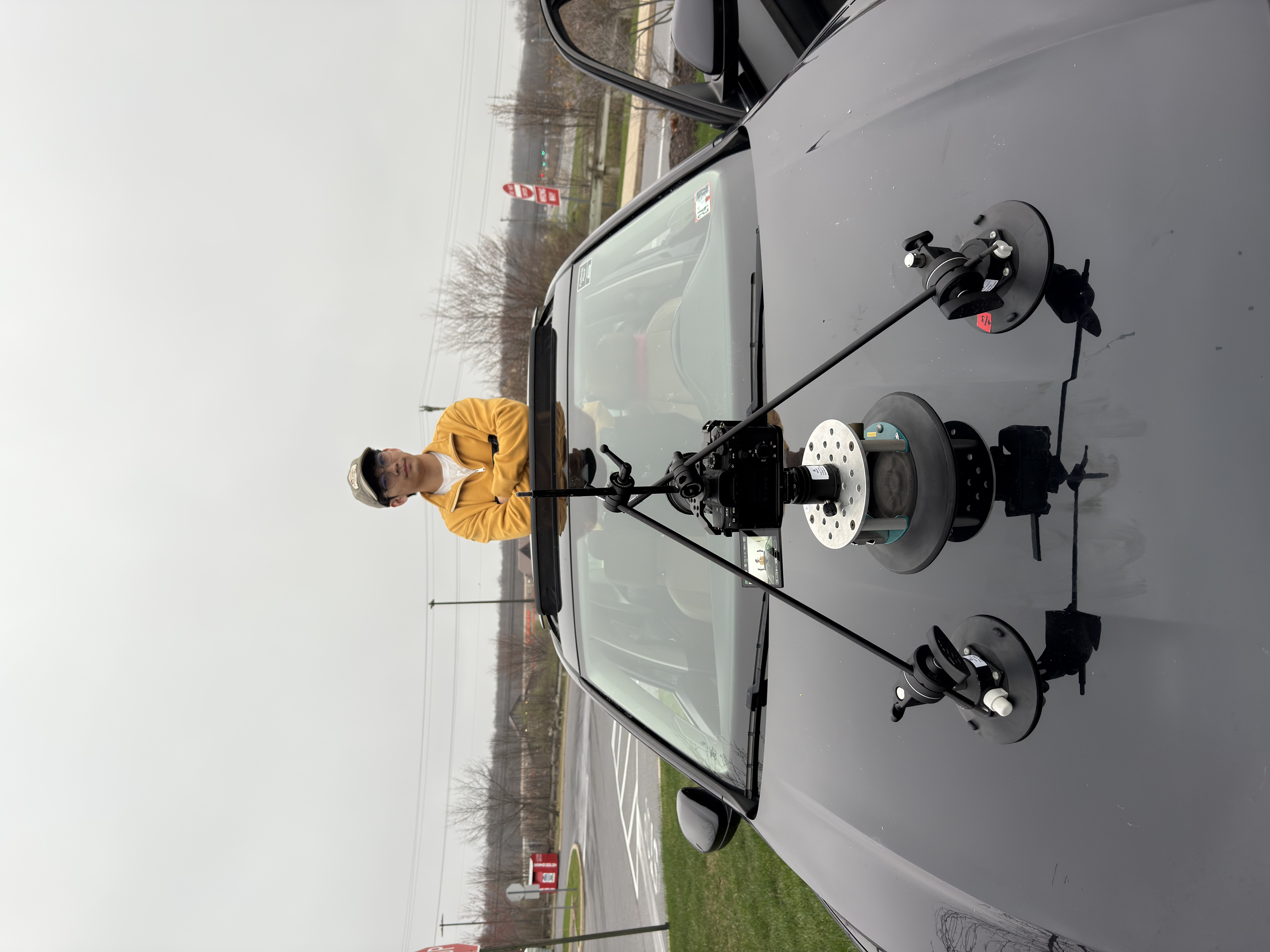 A camera rig is secured to a car hood with suction mounts while a person in a yellow jacket stands through the sunroof.