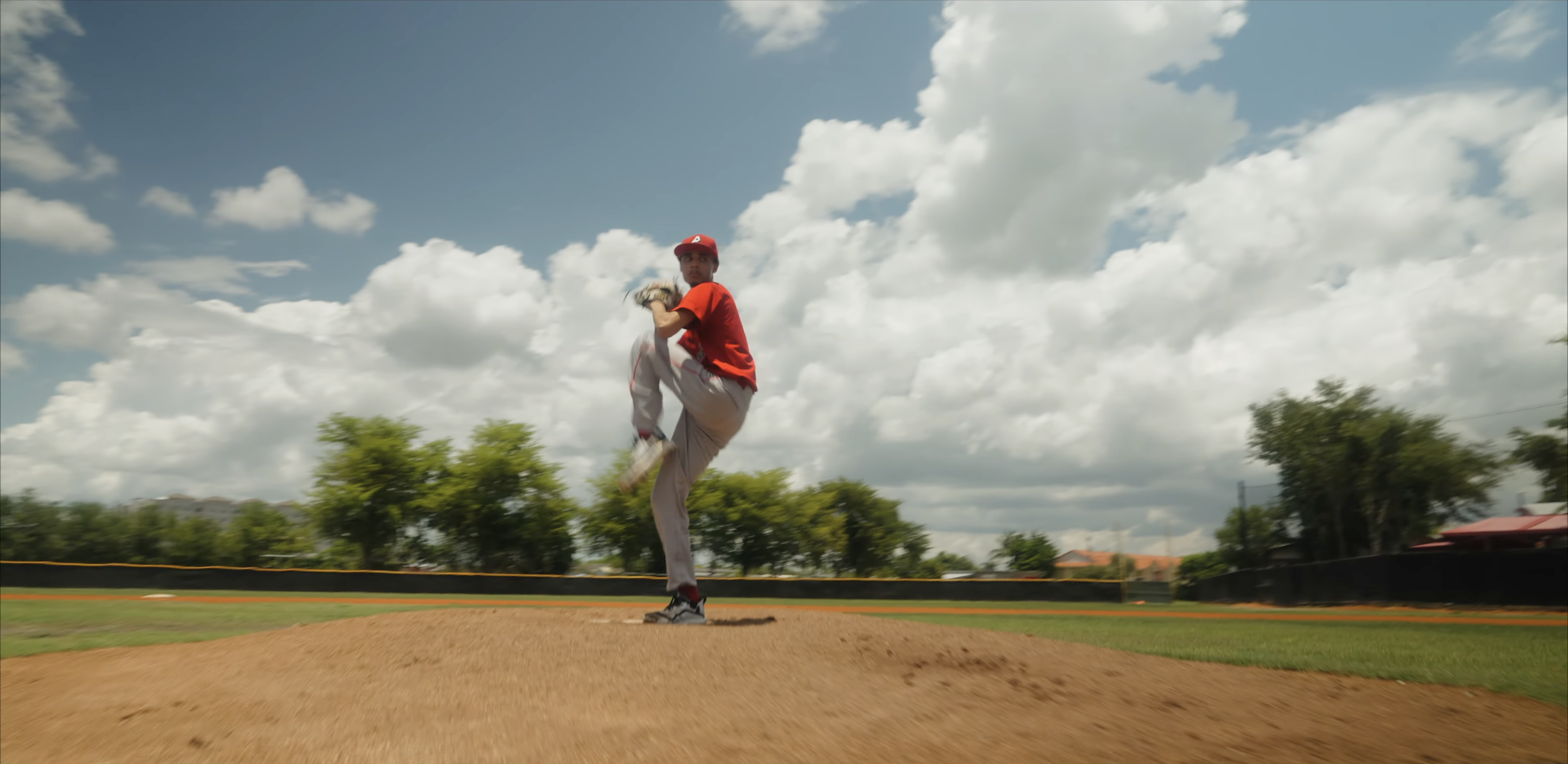 A person in a red shirt moves across a baseball field beneath towering white clouds.