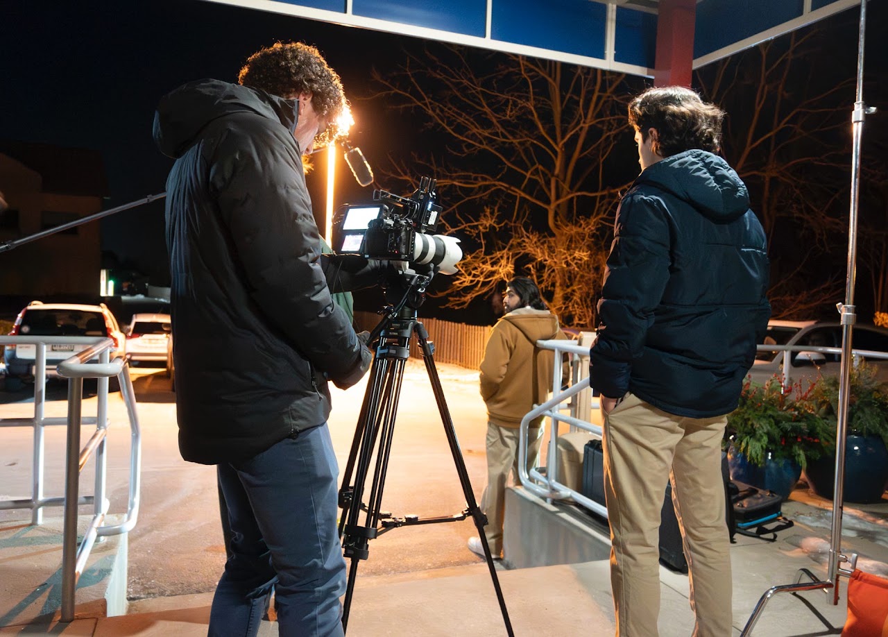 A cinematographer operates a camera on a tripod while filming a person standing outdoors at night.