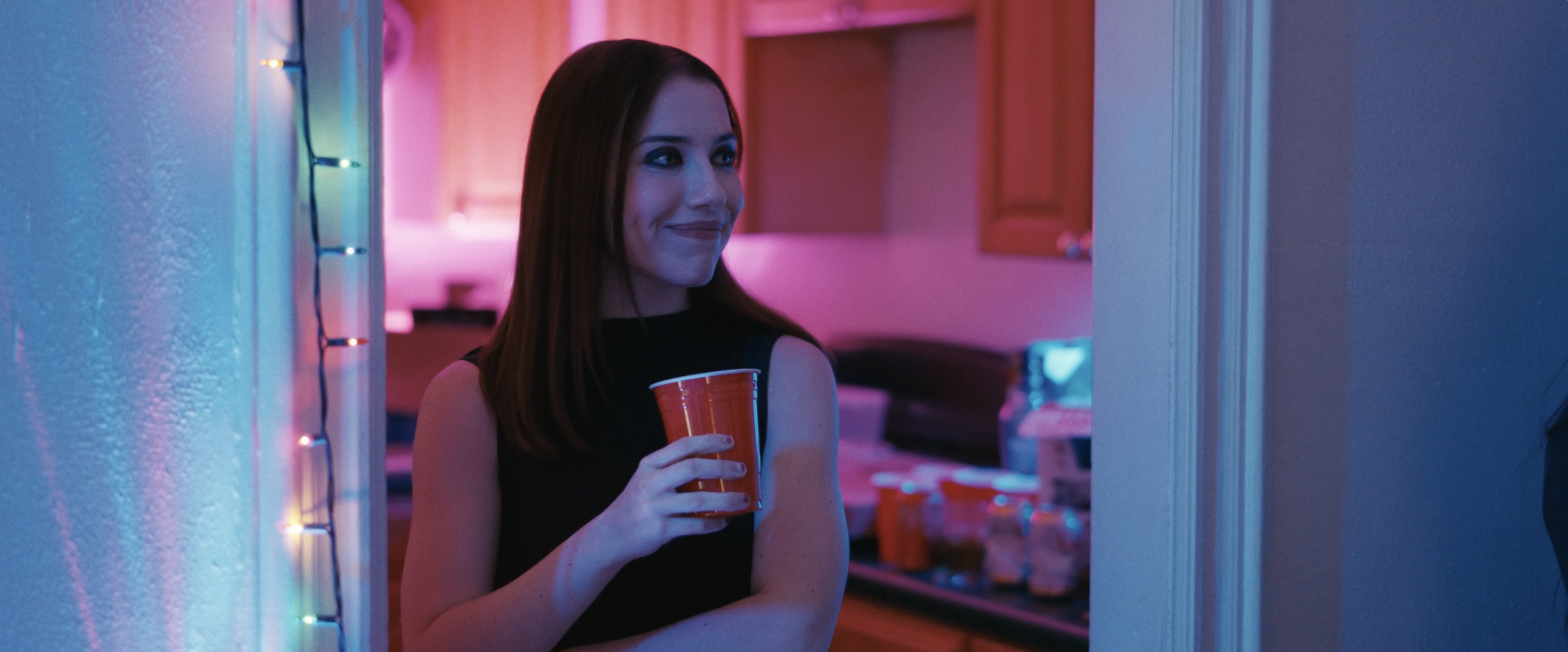 A young woman with long dark hair holds a red plastic cup while standing in a doorway illuminated by blue and pink neon lighting.