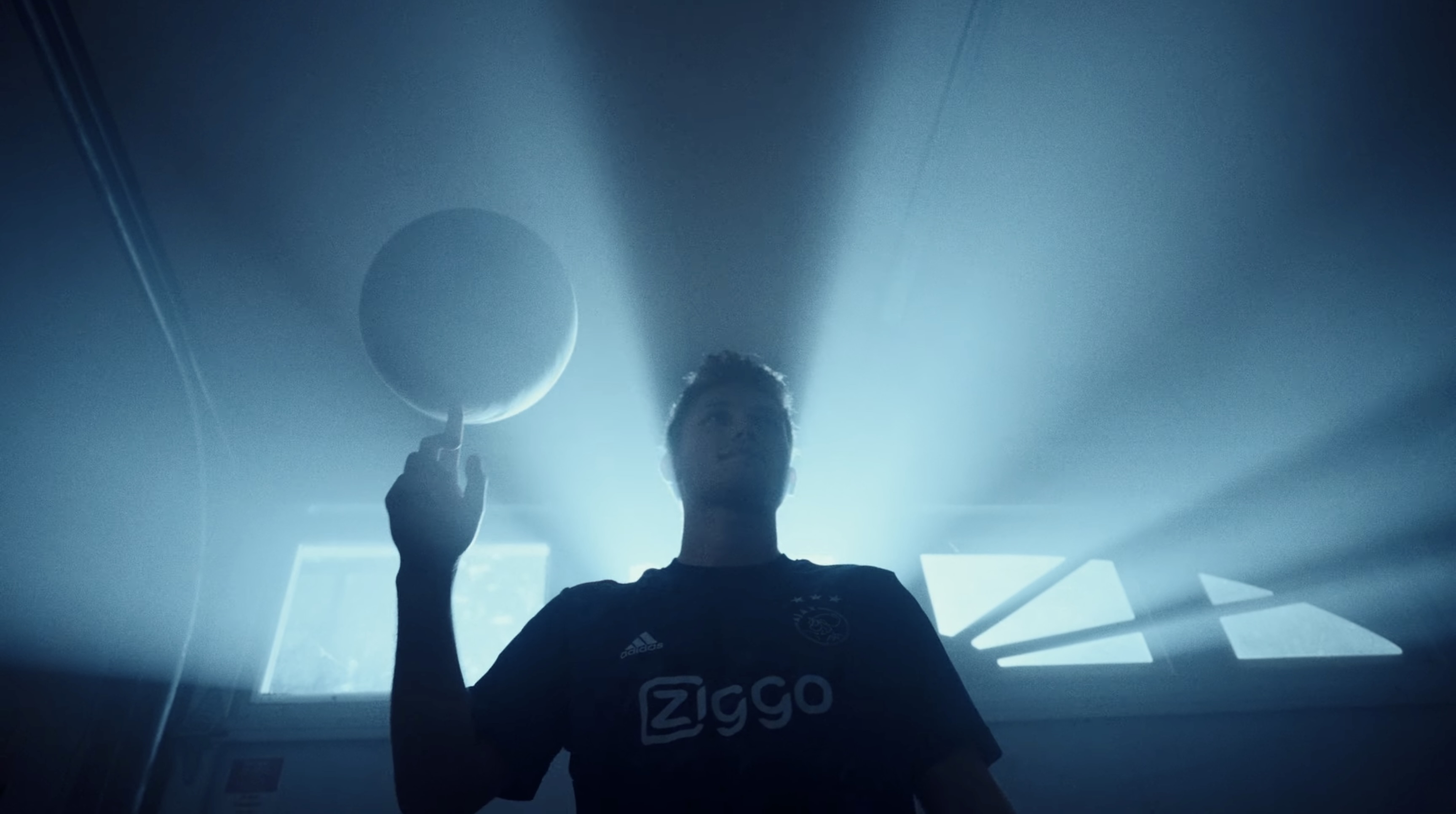 A silhouetted person raises a hand in blue fog beneath glowing geometric ceiling panels.
