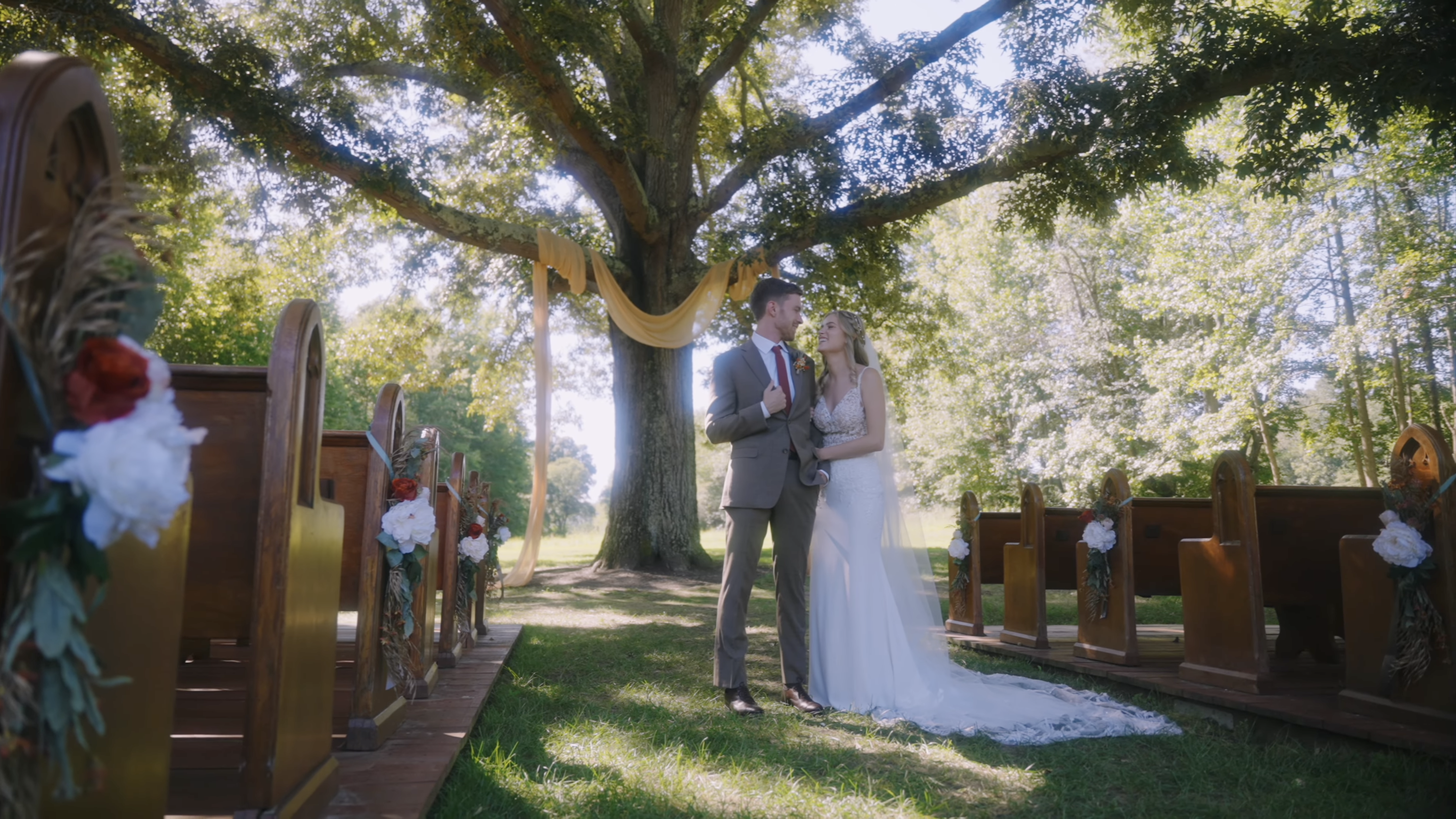 A bride and groom stand together under a large oak tree in an outdoor wedding ceremony setting with wooden pews arranged on either side.