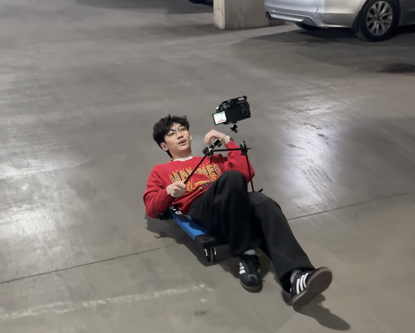 A camera operator lies on a low rolling platform while holding a mounted camera in a parking garage.