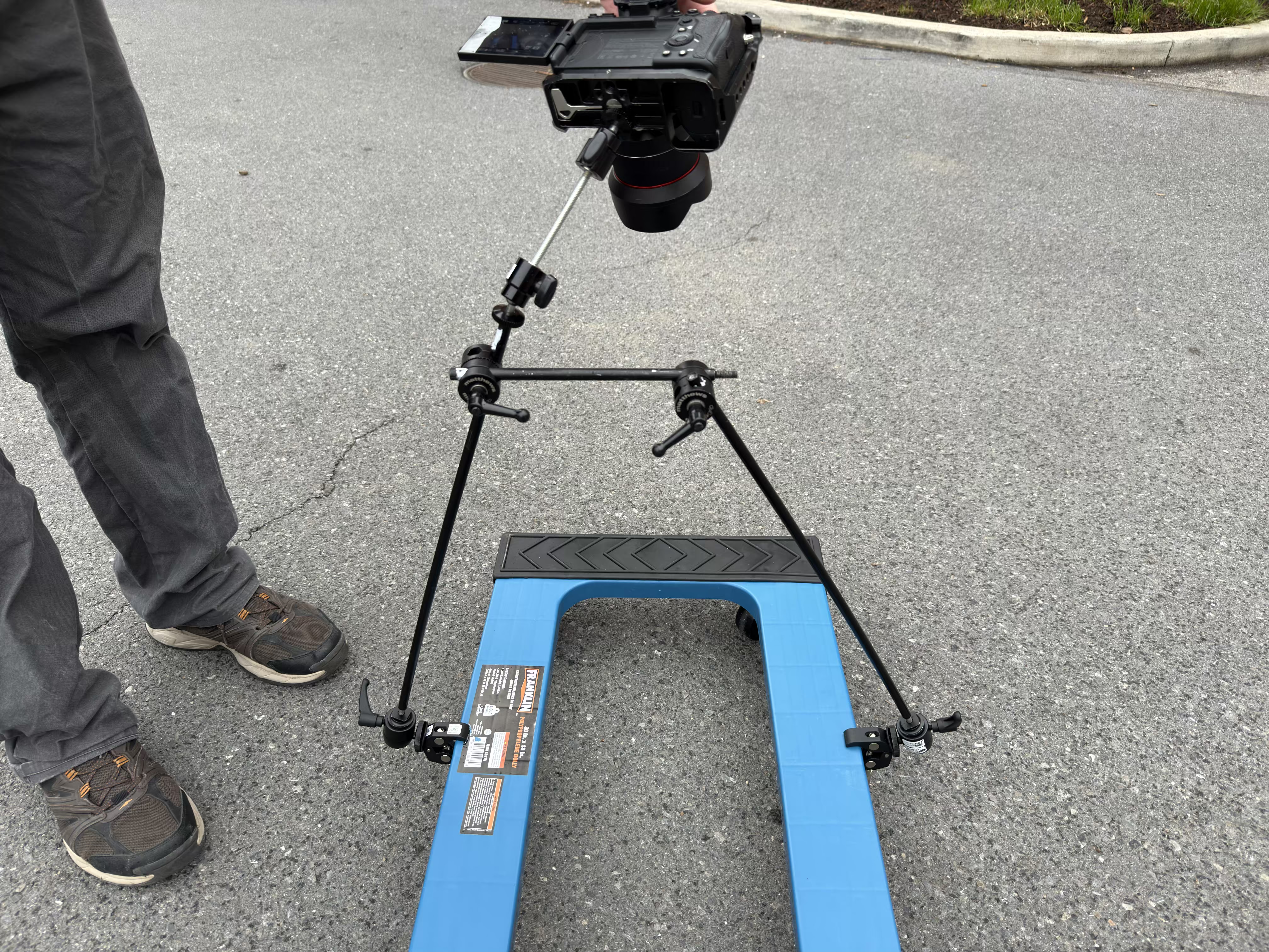 Close view of a camera mounted to a small blue wheeled rig on pavement beside a crew member.