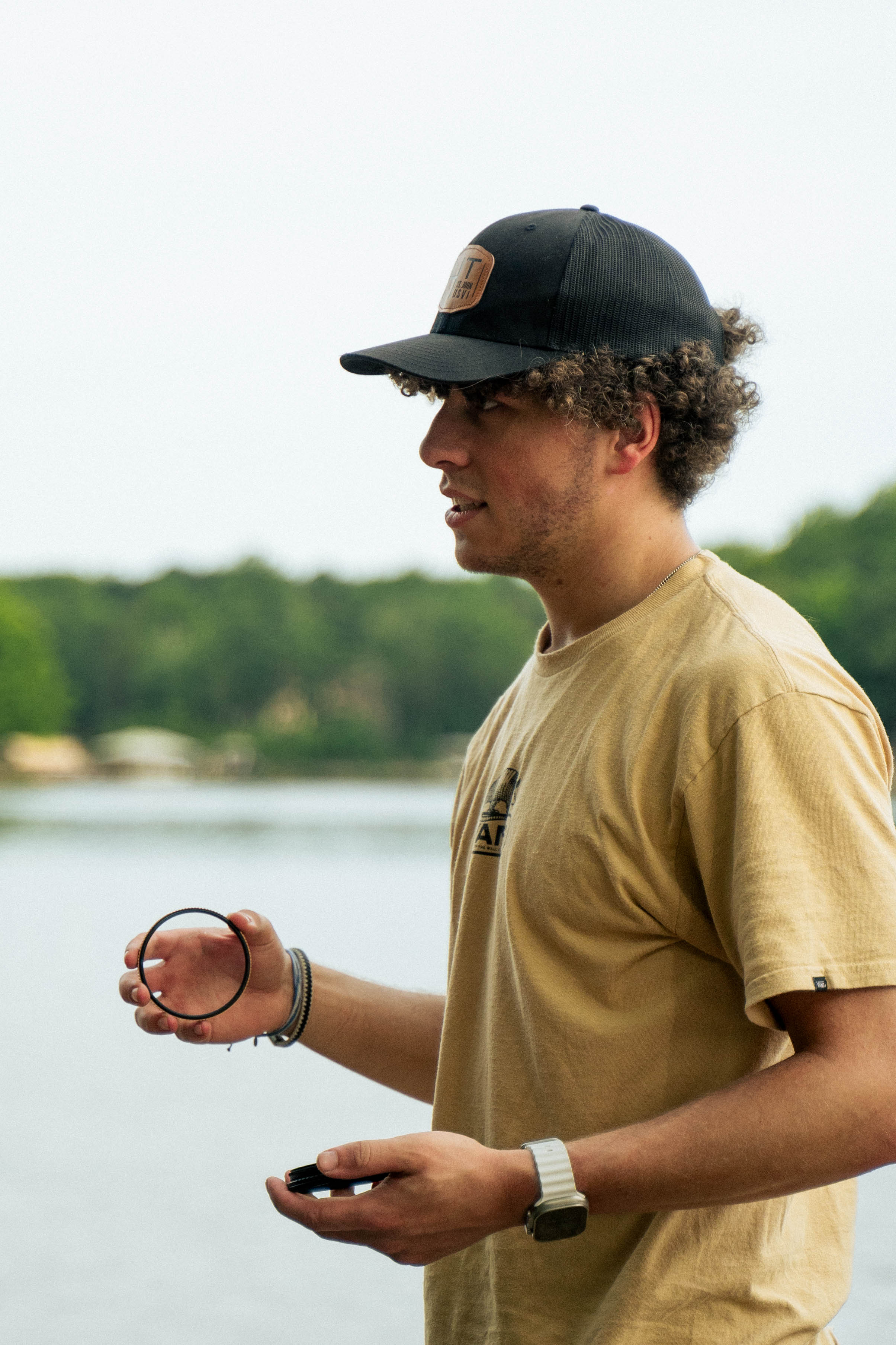 A young man with curly hair wearing a baseball cap and tan t-shirt holds a small black ring in one hand and a small black object in the other while standing outdoors.