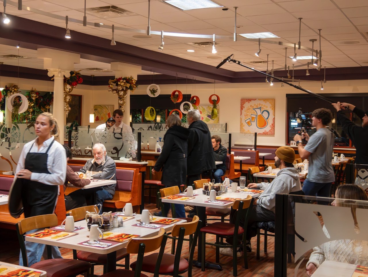 A film crew member holds a boom microphone over a busy restaurant scene while a server walks past tables of diners.