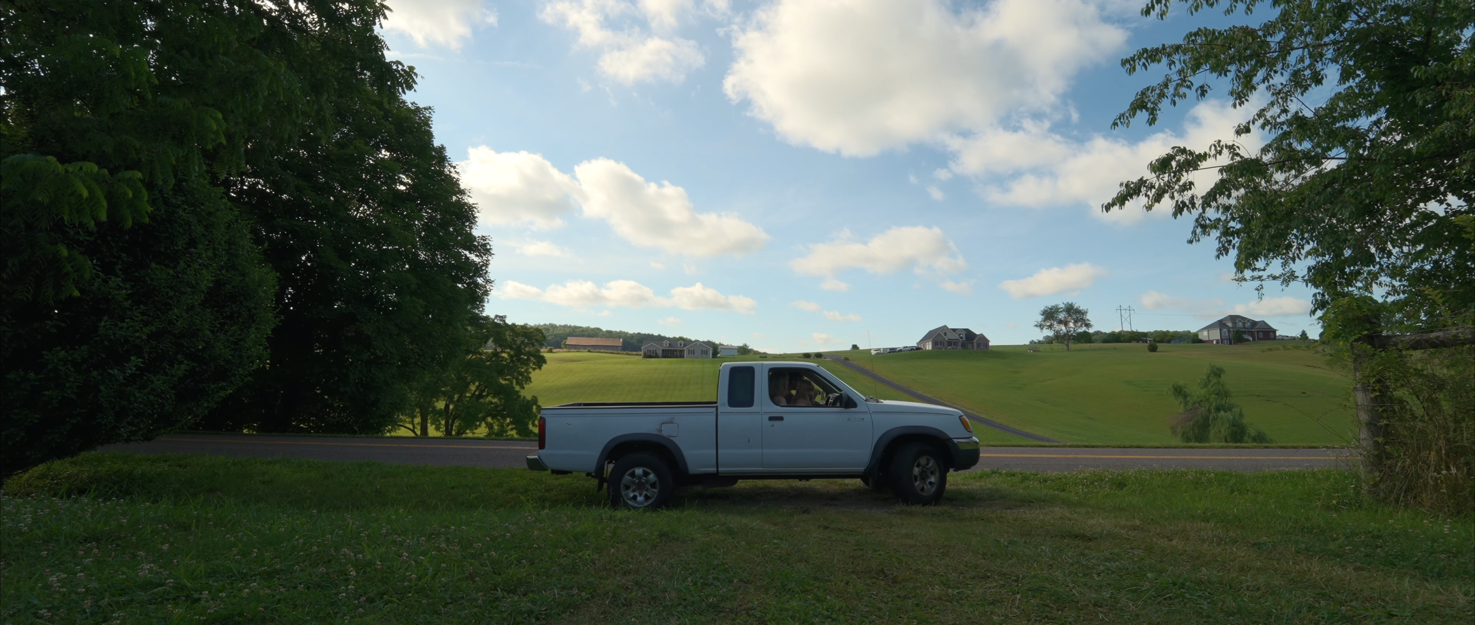 A light-colored pickup truck parked on a grassy roadside with a rolling green landscape and houses in the background under a blue sky with clouds.