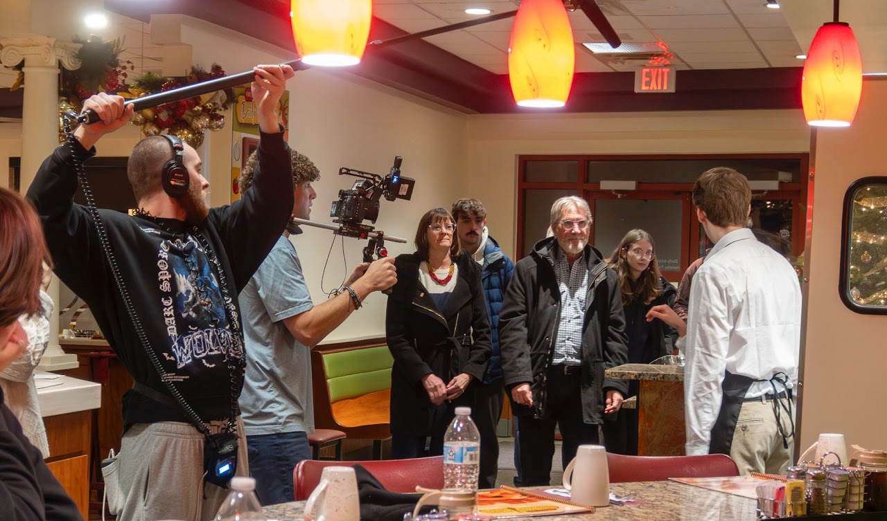 A film crew records a scene in a restaurant with a boom operator holding a microphone overhead and a camera operator filming a person in a white shirt.