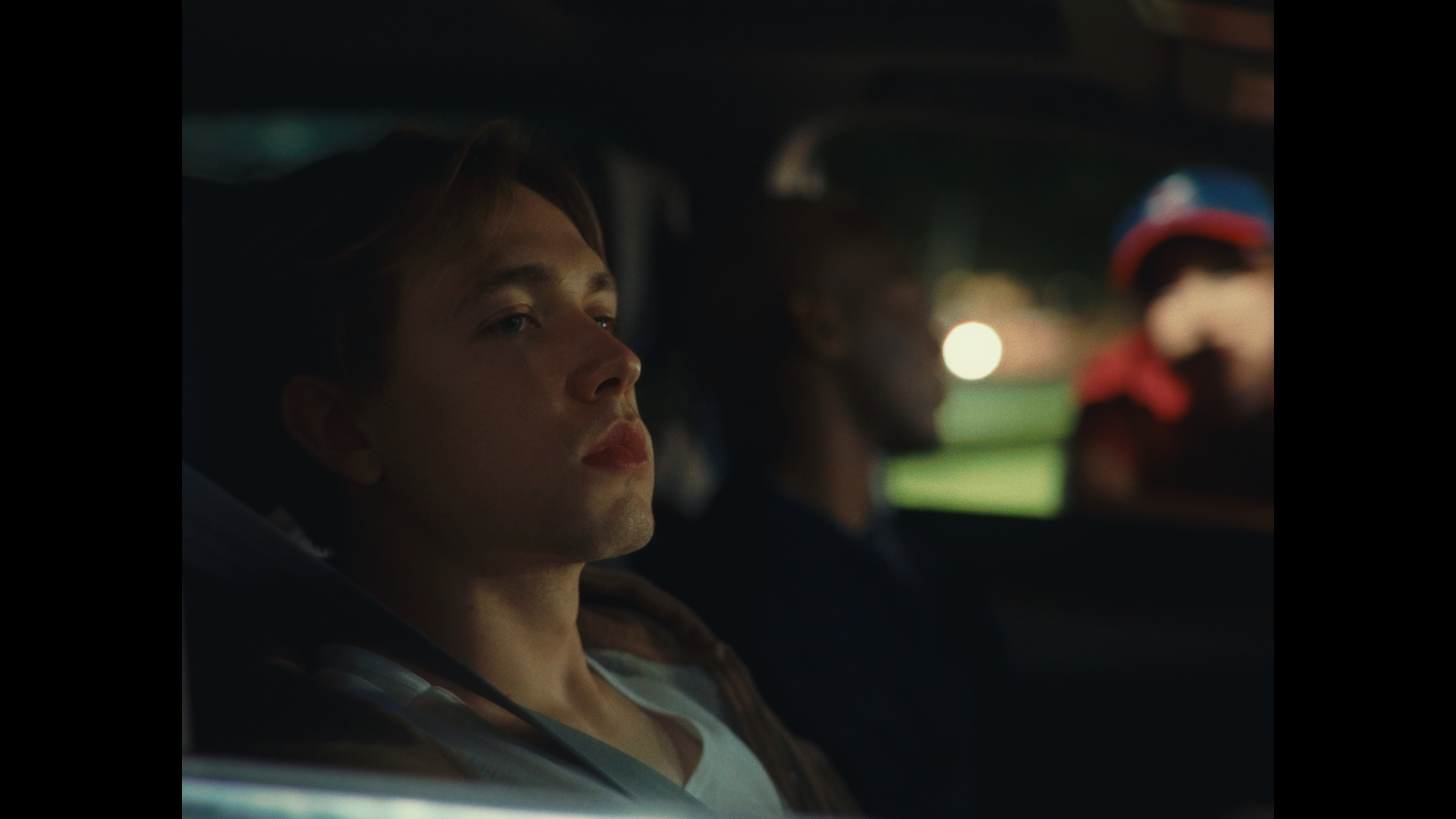 A close-up shot of a young man with light hair sitting in the passenger seat of a car at night, looking away from the camera.