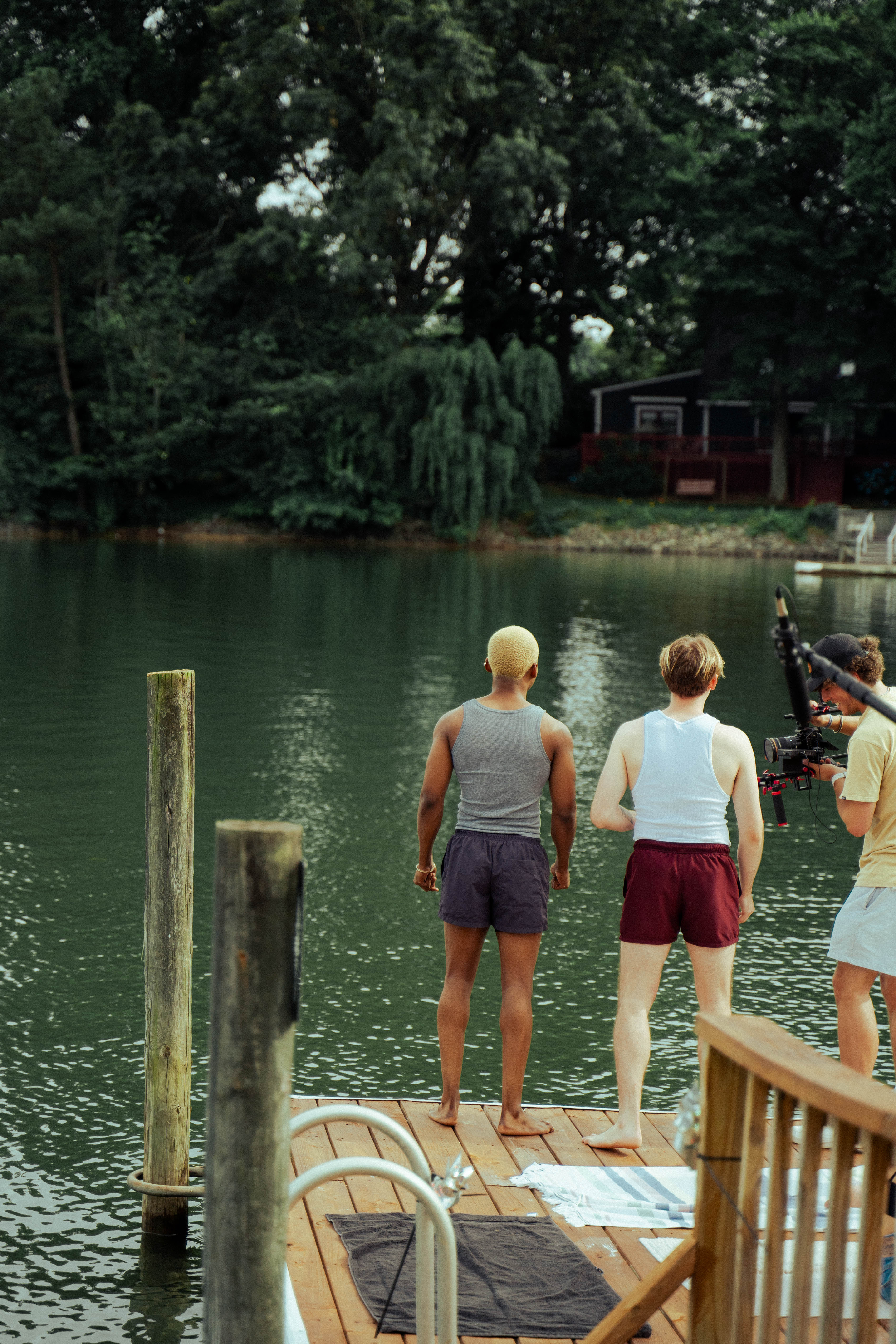 Two men stand on a wooden dock overlooking a lake while a camera operator films them from the side.