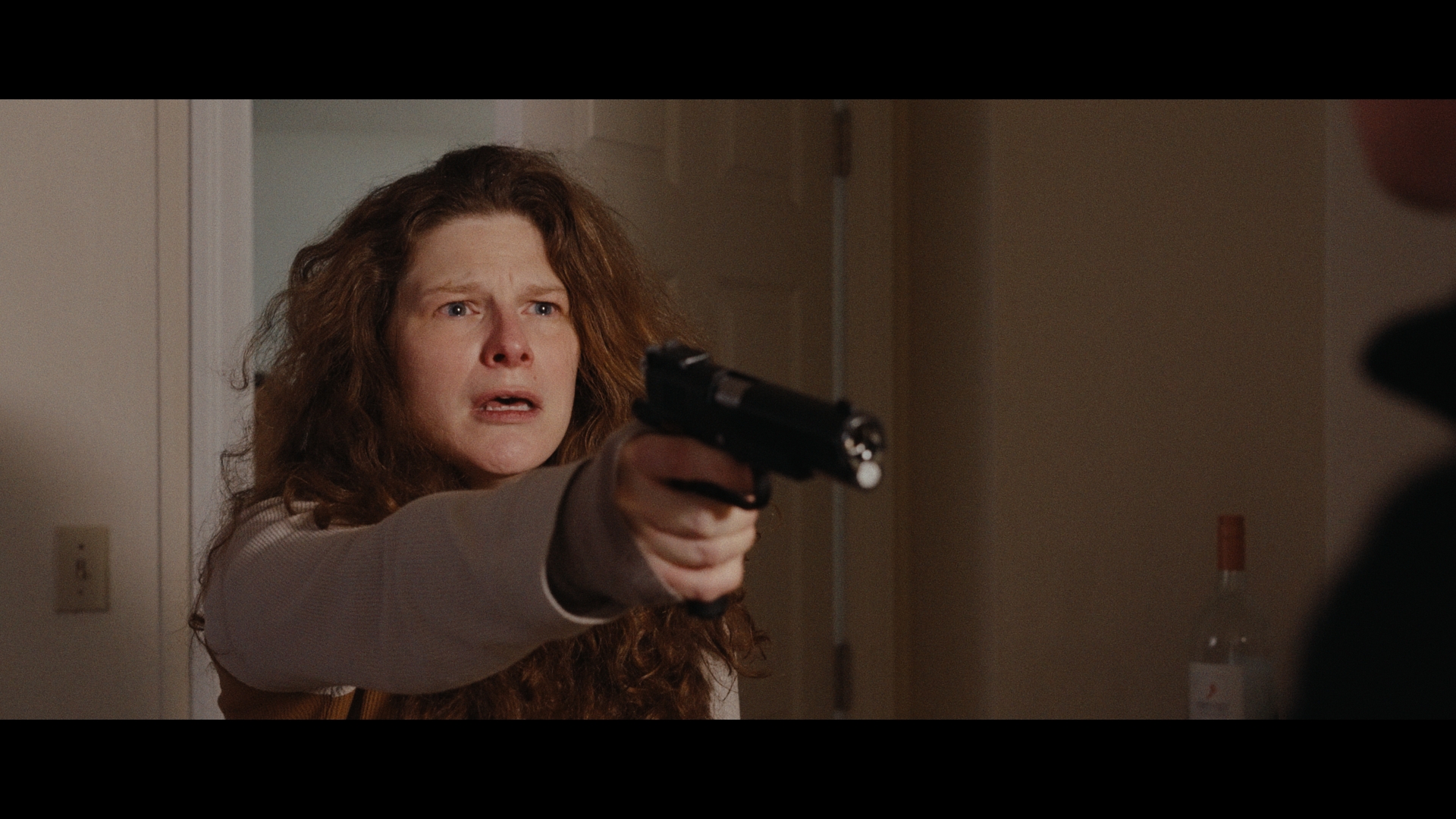 A woman with long curly hair holds a black handgun pointed forward with a tense expression.