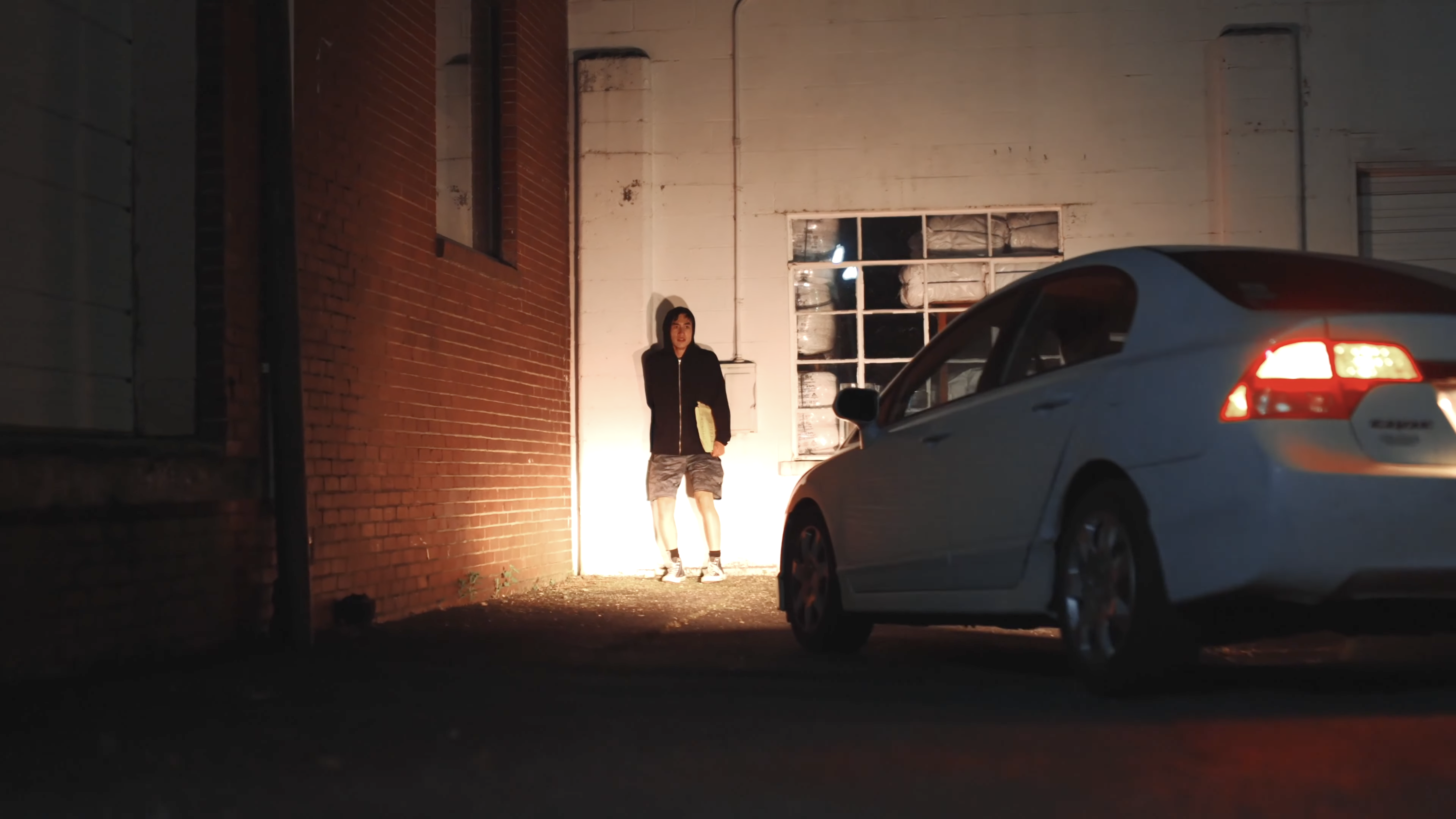 A person in a dark hoodie and shorts stands against a white wall at night, illuminated by the headlights of a white car parked in the foreground.