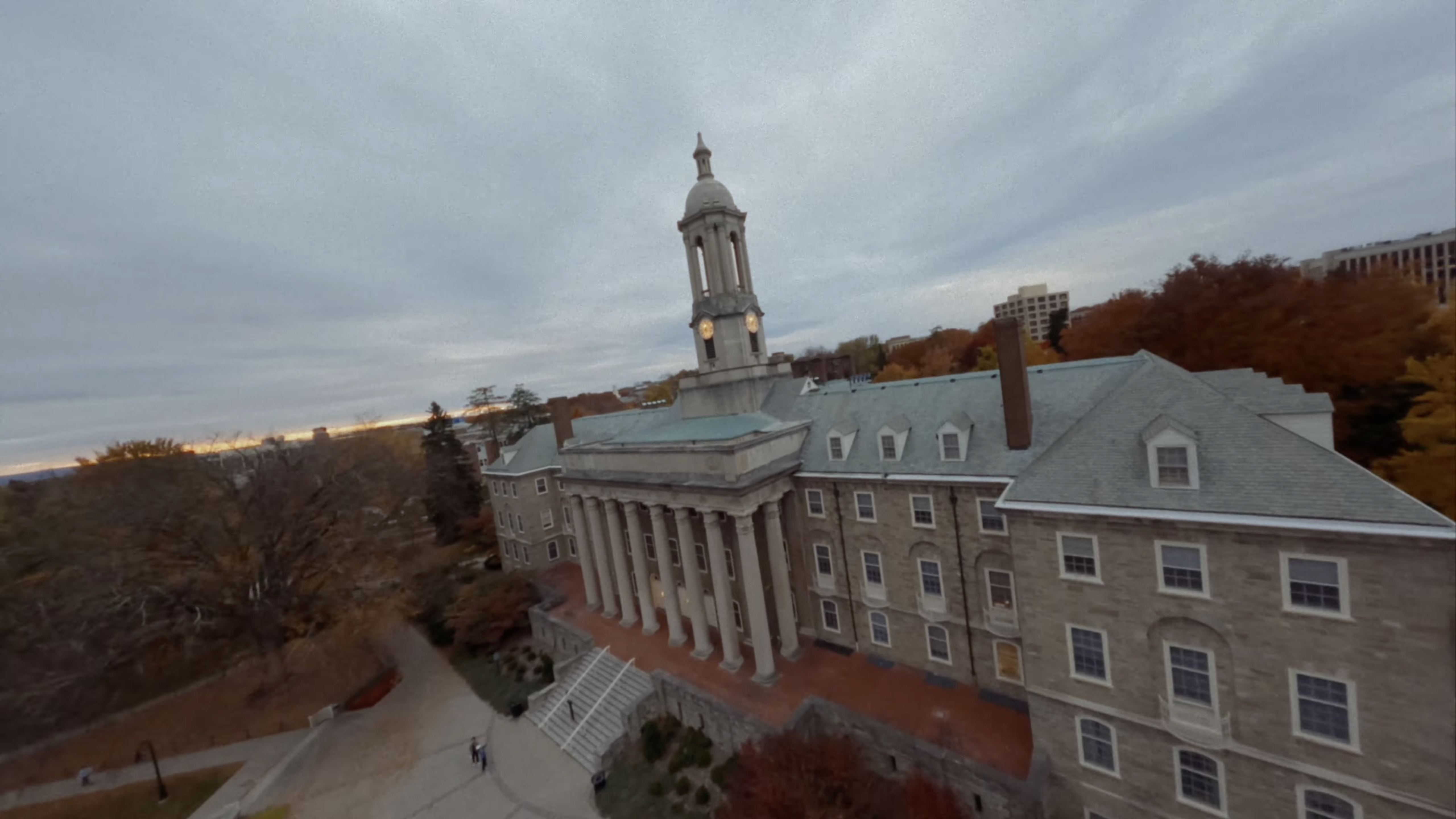 An aerial view of a large stone building with a central clock tower and columns, surrounded by autumn trees under a cloudy sky.