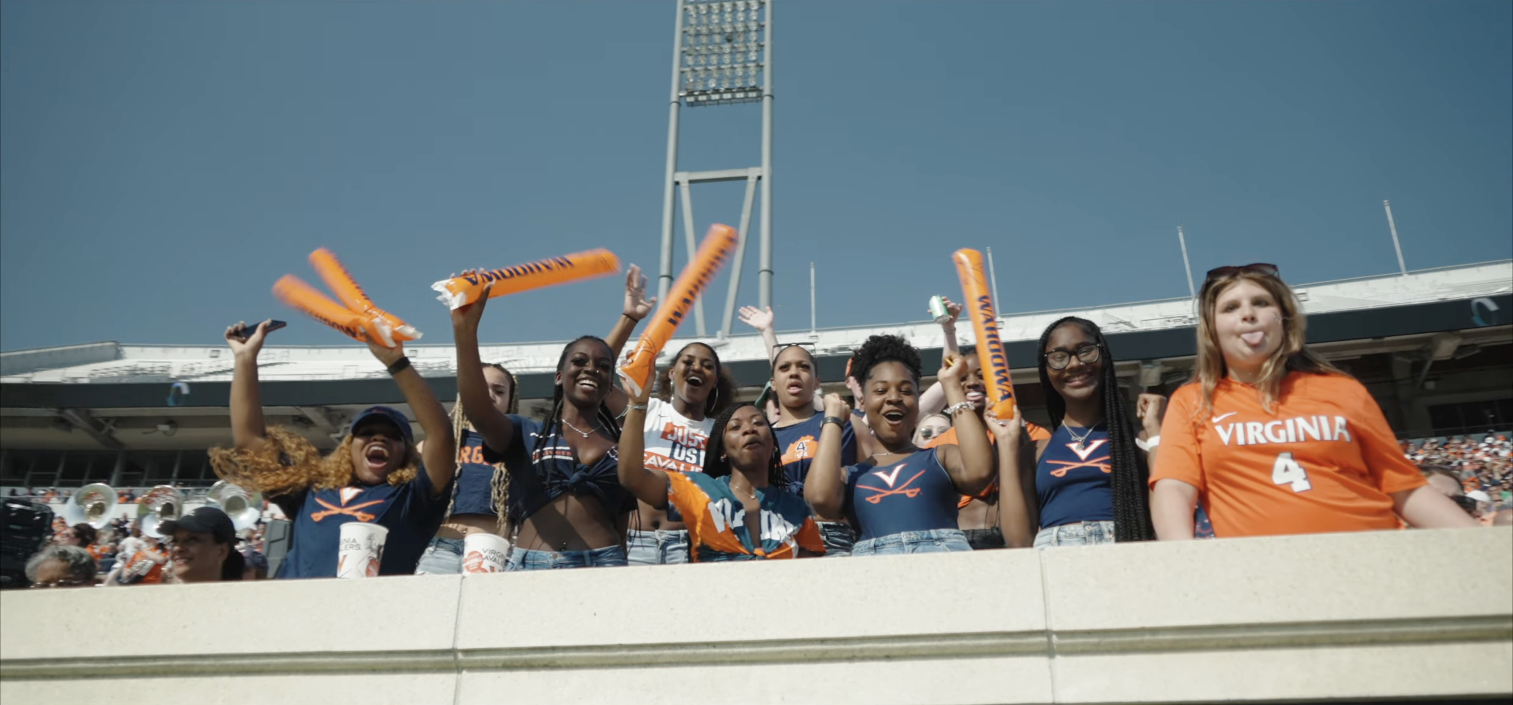 A group of young women in a stadium cheering with orange inflatable sticks, wearing Virginia Cavaliers apparel under a clear blue sky.